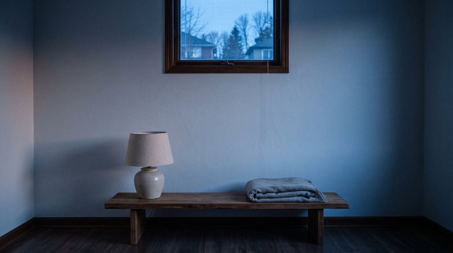 An unlit lamp and a folded throw on a low bench in a bedroom at dusk, washed in cold blue light