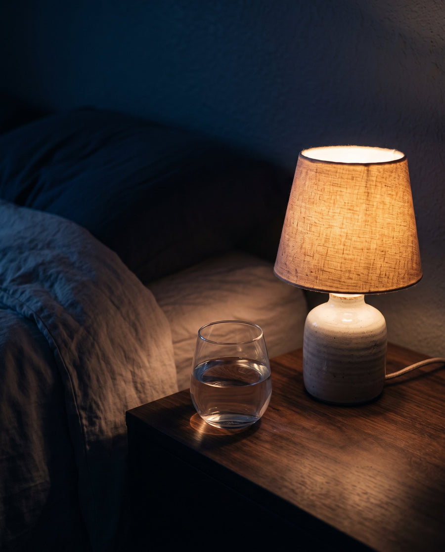 A glass of water and a softly lit lamp on a bedside table in a dark bedroom